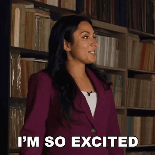 a woman in a purple jacket is standing in front of a bookshelf and says i 'm so excited