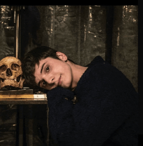 a young man in a blue sweater leans his head on his arm next to a skull