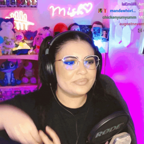 a woman wearing headphones and glasses is holding a rode microphone in front of a neon sign that says misky