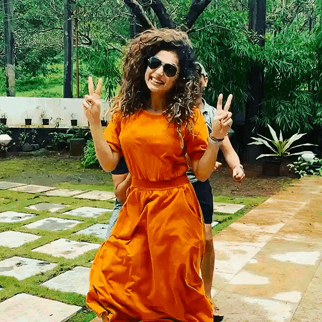 a woman wearing an orange dress and sunglasses giving a peace sign