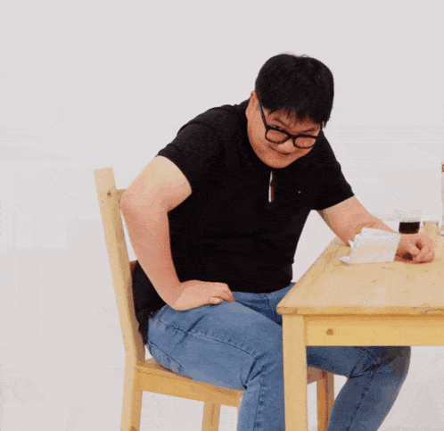 a man wearing glasses sits at a wooden table with his hands on his hips