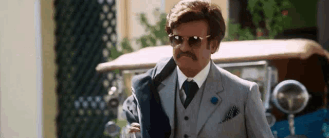 a man in a suit and tie with a mustache and sunglasses is walking in front of a car .