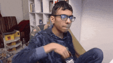 a young man wearing glasses and a blue sweatshirt sitting on a couch smoking a cigarette