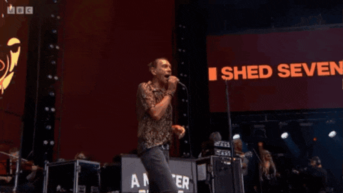 a man singing into a microphone on a stage with a bbc logo in the corner