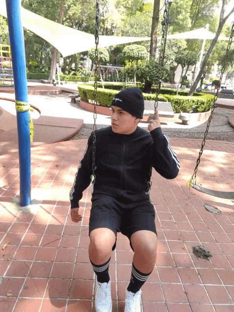a young man wearing a black beanie sits on a swing in a park
