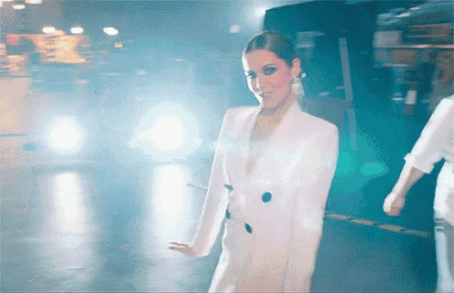 a woman in a white suit is dancing in front of a car