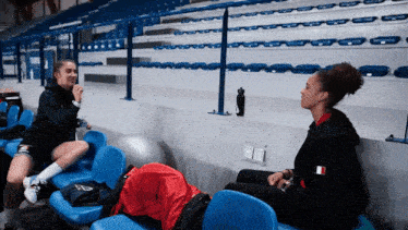 two women sit on blue seats in an empty stadium talking to each other