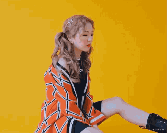 a woman wearing a plaid jacket and a black shirt is sitting on a yellow background
