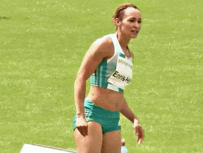 a female athlete wearing green shorts and a crop top is standing on a field with her hands in the air .