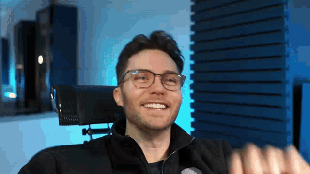 a man wearing glasses and a black jacket smiles while sitting in a chair