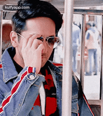 a man wearing sunglasses and a denim jacket is sitting on a bus and adjusting his glasses .