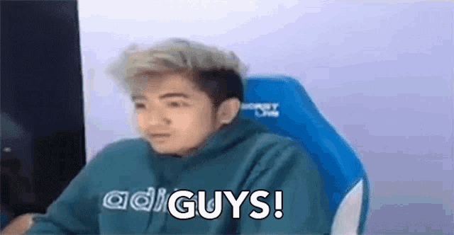 a young man in a green hoodie is sitting in a blue chair and saying `` guys '' .