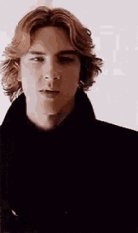a man with long red hair is wearing a black coat and looking at the camera .