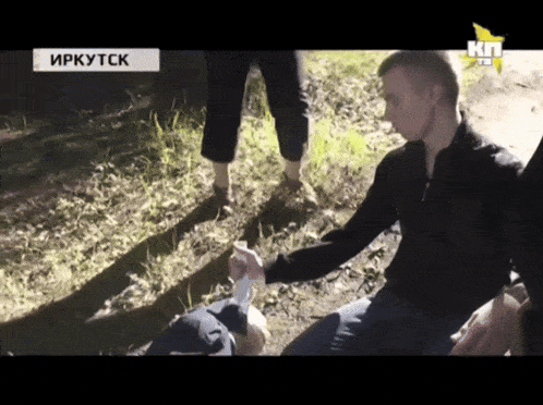 a man in a black jacket is kneeling down in the grass with a kn tv logo in the corner