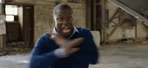 a man in a blue sweater is standing in front of a car with the hood open and screaming .