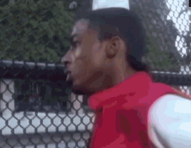 a man wearing a red vest and a white hat is standing in front of a chain link fence .