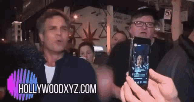 a man is taking a picture of another man with a cell phone with hollywoodxyz.com written on the bottom
