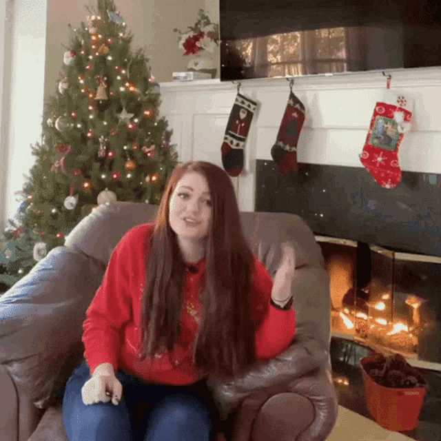 a woman in a red sweater sits in front of a christmas tree and fireplace