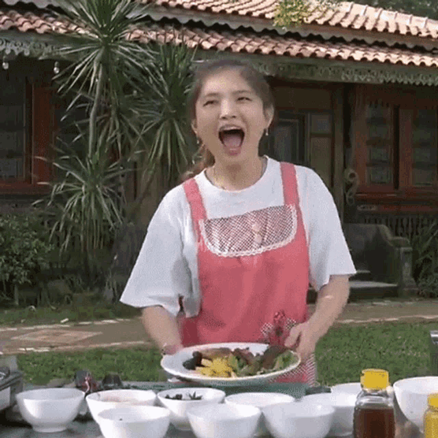 a woman in a pink apron is holding a plate of food in front of a house .