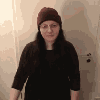 a woman wearing glasses and a beanie stands in front of a white door