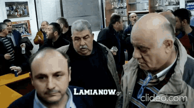 a group of men are gathered in a room with the website lamianow.gr in the background