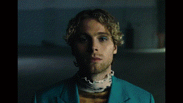 a young man with curly hair and a beard is wearing a necklace and a white shirt .