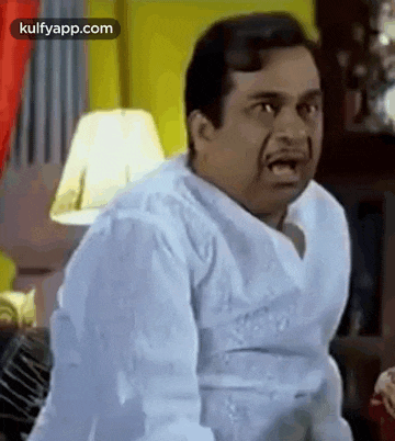 a man in a white shirt is making a funny face while sitting on a couch in a living room .