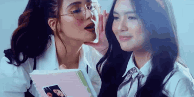 two girls are whispering into each other 's ear while holding a book .