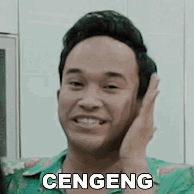 a man in a green shirt is smiling and holding his hand to his ear with the word cengeng on it .