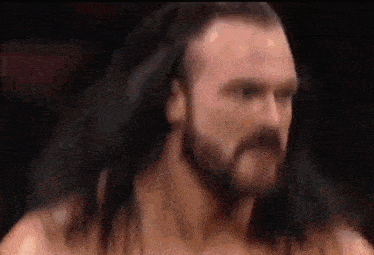 a wrestler with long hair and a beard is standing in a ring .