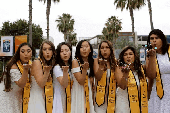 a group of women wearing yellow sashes that say class of 2019 blowing confetti in the air