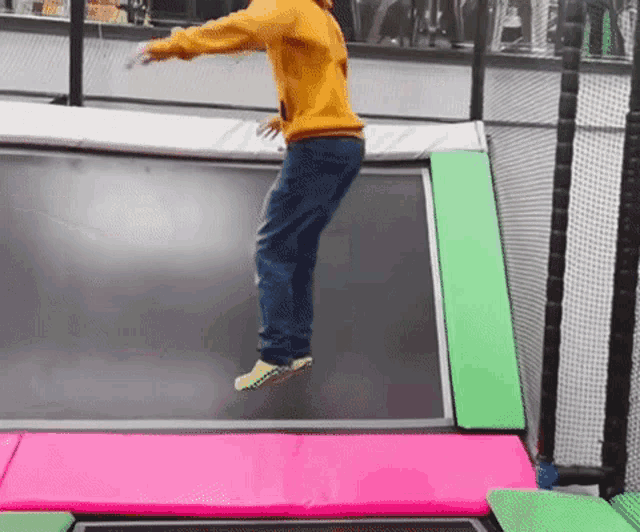 a person in a yellow sweatshirt is jumping on a pink and green trampoline