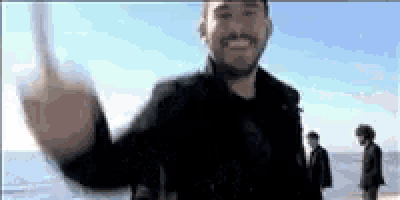 a man with a beard is waving his hand at the camera while standing on a beach .