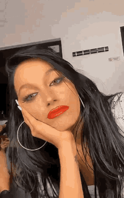 a woman with red lipstick and hoop earrings is sitting with her hand on her face .