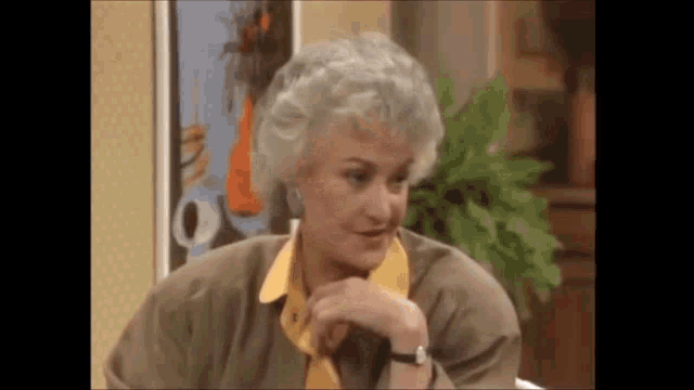 a woman with gray hair and a yellow collar is sitting at a table with her hand on her chin .