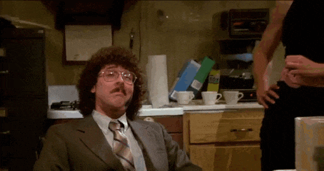 a man with curly hair and glasses is sitting in a kitchen with a bag of gas in the background