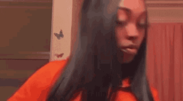 a woman with long black hair is wearing an orange shirt and standing in a room .