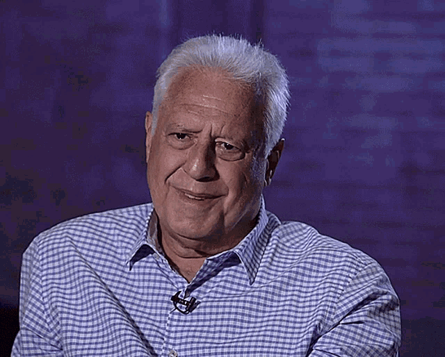 an older man wearing a blue and white checkered shirt with a microphone on his neck