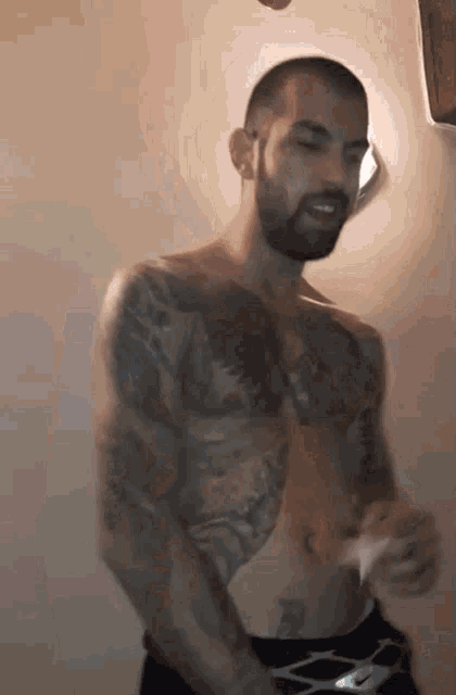 a shirtless man with a beard and tattoos is standing in front of a wall .