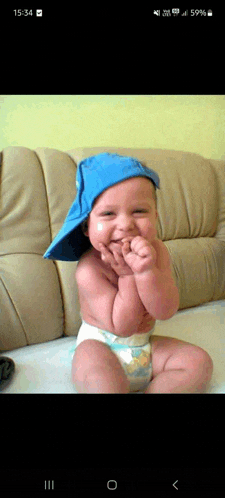 a baby wearing a diaper and a blue hat is sitting on a couch at 15:34