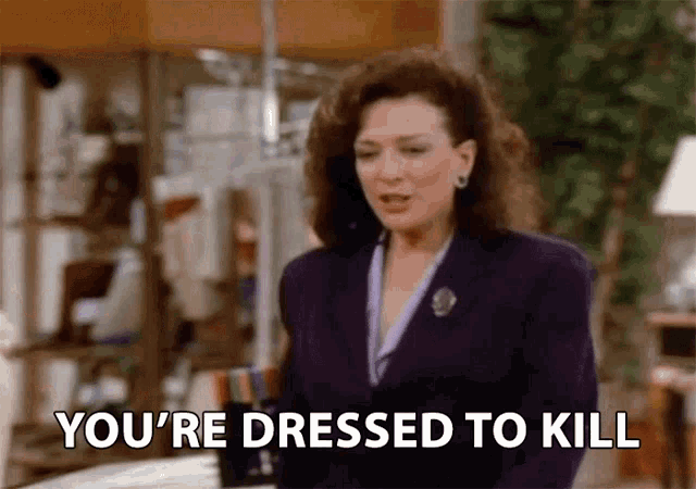 a woman in a purple suit is standing in a room and says you 're dressed to kill .