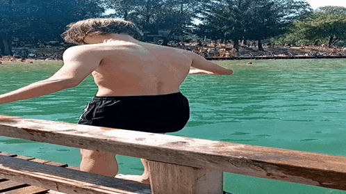 a shirtless man in black shorts is standing on a wooden dock overlooking a body of water