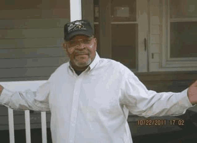 a man in a white shirt and black hat is standing in front of a house .