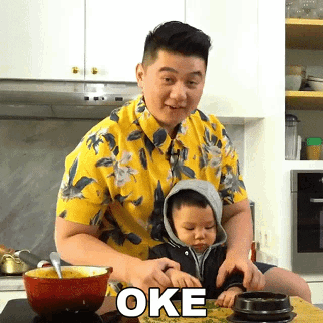 a man in a yellow shirt is playing with a baby in a kitchen and the word oke is above them