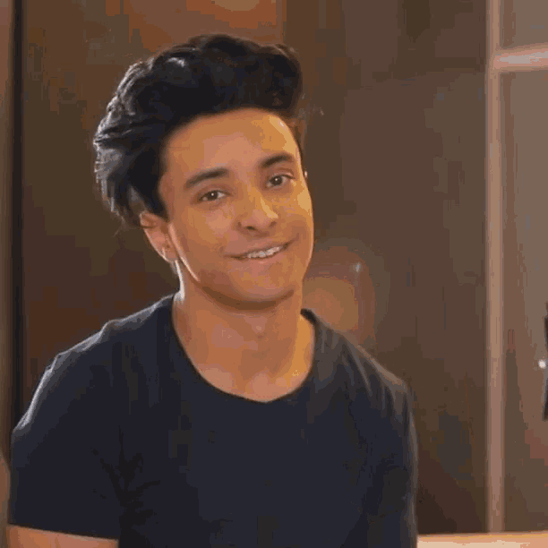 a young man in a black t-shirt is smiling and waving at the camera .