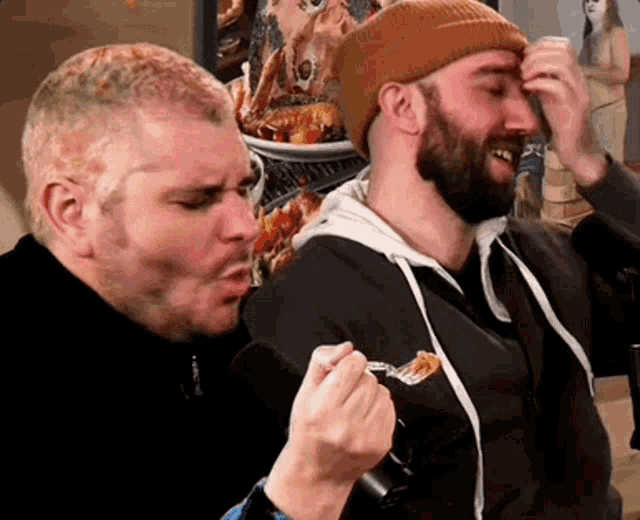 two men are sitting next to each other eating food . one of the men is holding a fork in his hand .