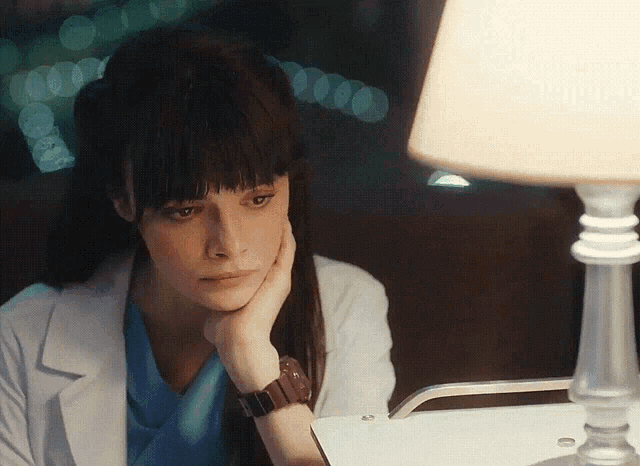 a woman in a lab coat sits at a table with her hand on her chin