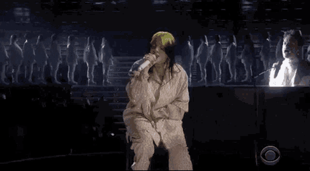 billie eilish is singing into a microphone while sitting on a stage in front of a piano .
