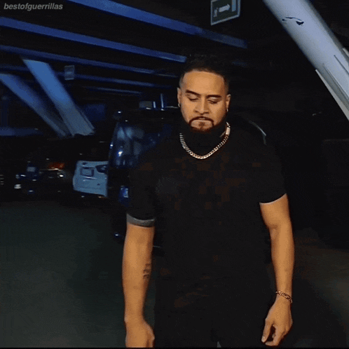 a man with a beard wearing a black shirt and a gold chain stands in a parking garage
