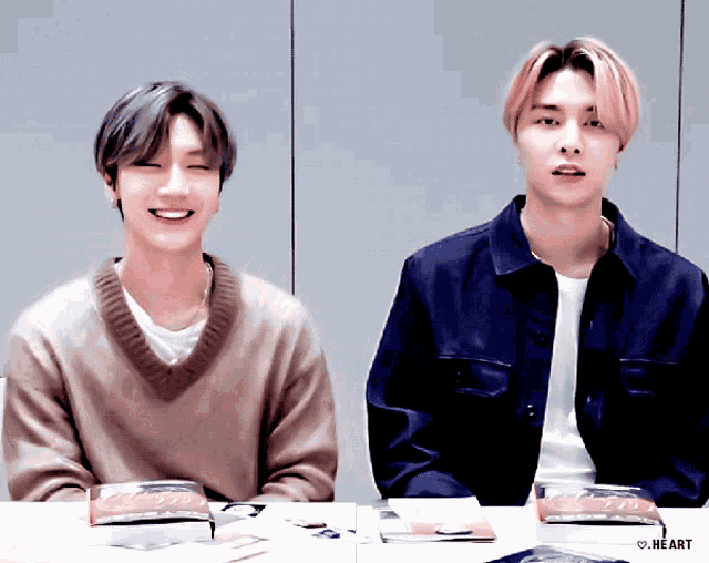 two young men are sitting at a table and one of them is wearing a brown sweater and a blue jacket